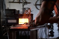 Back view of anonymous tattooed young male forger in apron heating metal in furnace while working in grungy smithy — Stock Photo