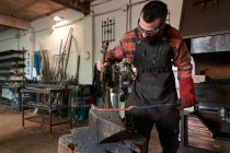 Concentrated young bearded blacksmith in apron and protective goggles striking heated metal with hammer on anvil while working in forge — Stock Photo