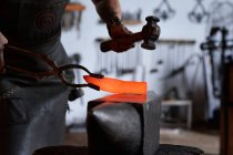 Vue latérale d'un jeune faussaire tatoué anonyme dans un tablier chauffant du métal dans un four tout en travaillant dans une forge grunge — Photo de stock