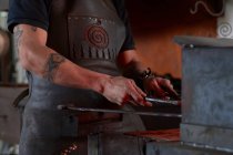 Vue latérale d'un jeune faussaire tatoué anonyme dans un tablier chauffant du métal dans un four tout en travaillant dans une forge grunge — Photo de stock