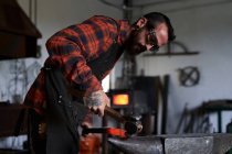 Side view of young brutal bearded craftsman in apron and goggles heating and striking iron on anvil with hammer during work in traditional smithy — Stock Photo