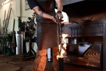 Crop anonymous tattooed male blacksmith in casual clothes and apron heating metal pliers in flame during forging process in workshop — Stock Photo