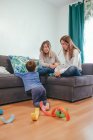 Full length cheerful young lesbian couple in casual wear resting on cozy couch and looking at cute son playing on floor — Stock Photo
