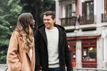 Allegro giovane coppia indossa vestiti caldi che si tengono per mano e si guardano con sorrisi mentre camminano insieme sul marciapiede di asfalto nella città moderna — Foto stock