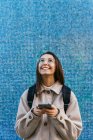 Урожай привлекательный улыбающийся студентка в теплой одежде с рюкзаком с помощью современного мобильного телефона на тротуаре на улице — стоковое фото