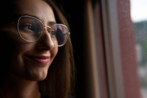 Crop positive junge Frau mit dunklen Haaren trägt eine Brille steht in dunklen Raum und schaut aus dem Fenster mit Lächeln — Stockfoto