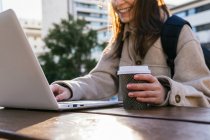 Unrecognizable happy smiling female student in warm coat and eyeglasses with backpack working on home assignment on netbook while sitting at table with takeaway coffee in sunny park — Stock Photo
