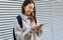 Crop joyful young female in warm clothes and eyeglasses with backpack browsing contemporary mobile phone while standing on sidewalk — Stock Photo