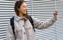 Joyeux jeune femme en tenue chaude avec sac à dos prenant selfie sur téléphone portable tout en se tenant sur le trottoir — Photo de stock