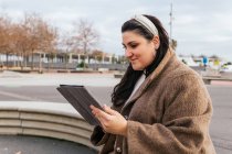 Young smiling plump female in casual wear browsing internet on tablet while resting on bench against urban building in fall — Stock Photo