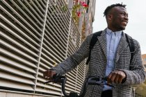 Young content African American male employee in coat with bike standing on urban pavement against ribbed wall and looking away — Stock Photo