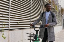 Young content Empleado afroamericano en abrigo con bicicleta parado en pavimento urbano contra pared acanalada y mirando hacia otro lado - foto de stock