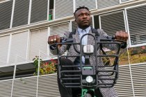 Jovem conteúdo afro-americano masculino empregado no casaco com bicicleta de pé no pavimento urbano contra a parede com nervuras — Fotografia de Stock