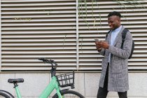 Bajo ángulo de contenido Gerente masculino afroamericano escribiendo mensajes de texto en el teléfono celular cerca de la bicicleta en la ciudad - foto de stock