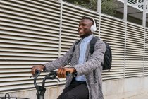Young happy ethnic male office employee listening to music in headset while riding bicycle on city street and looking forward — Stock Photo