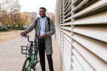 Junge zufriedene afroamerikanische männliche Angestellte im Mantel mit Fahrrad steht auf städtischem Bürgersteig vor gerippter Wand und blickt in die Kamera — Stockfoto