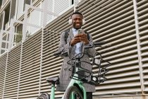 Low angle of content African American male manager in headset text messaging on cellphone near bicycle in town — Stock Photo