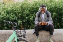 Young positive ethnic male office employee in headphones chatting on cellphone while resting near bicycle in town — Stock Photo
