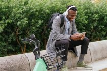 Молодой позитивный работник офиса этнического мужчины в наушниках болтает по мобильному телефону во время отдыха возле велосипеда в городе — стоковое фото