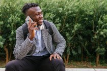 Jovem trabalhador de escritório afro-americano feliz com mochila falando no celular enquanto sentado contra o edifício urbano e olhando para longe — Fotografia de Stock