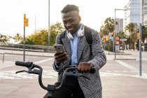 Joven alegre oficinista negro con auriculares charlando en el teléfono celular mientras está sentado en bicicleta en el pavimento urbano - foto de stock