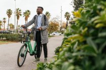 Ground level of young content African American male office worker with headphones on bike looking away between plants and trees in town — Stock Photo