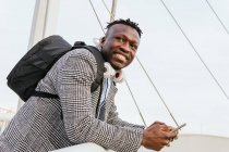 Side view of young cheerful African American male manager with backpack and cellphone leaning on fence of urban bridge while looking away — Stock Photo