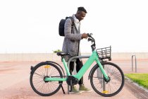 Vista lateral do funcionário do escritório masculino afro-americano usando o aplicativo no celular enquanto estaciona de bicicleta na cidade — Fotografia de Stock