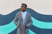 Exécutif masculin afro-américain réfléchi méconnaissable dans un masque debout avec les mains dans des poches près d'un mur lumineux et regardant loin en ville — Photo de stock