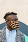 Анонімний споглядальний етнічний чоловічий менеджер в декоративній масці з мобільним телефоном, який дивиться далеко поблизу міської стіни під час пандемії COVID 19 — стокове фото