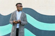 Anônimo contemplativa gerente masculino étnico em máscara ornamental com celular olhando para longe perto da parede urbana durante a pandemia COVID 19 — Fotografia de Stock