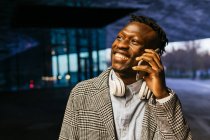 Joven trabajador de oficina masculino étnico sonriente en auriculares que hablan por teléfono celular mientras mira hacia otro lado en la calle por la noche - foto de stock