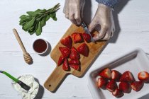 Top vista cultura chef irreconhecível em luvas de látex cortar morangos deliciosos frescos em tábua de corte de madeira na mesa perto de queijo creme — Fotografia de Stock