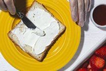 Top view crop anonymous female cook in latex gloves spreading yummy cream cheese on bread slice placed on yellow plate near cut strawberries — Stock Photo