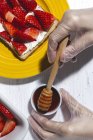 Overhead crop anonymous skilled female chef in gloves pouring sweet honey on delicious toast with cream cheese and cut strawberries while cooking in light kitchen — Stock Photo