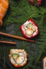 Savoureux sushi frais assorti servi sur des brindilles de plantes vertes sur une assiette noire avec sauce soja sur une table en marbre près de baguettes — Photo de stock
