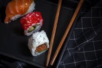 Верхний вид композиции вкусных свежих различных суши и бамбуковых палочек подается на черном блюде на клетчатой ткани — стоковое фото