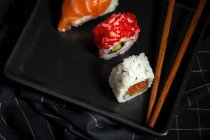 Composition vue du dessus de délicieux sushi frais divers et baguettes de bambou servis sur plateau noir sur tissu à carreaux — Photo de stock
