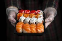 Crop chef irriconoscibile in guanti che mostra piatto con set di sushi assortiti appetibili in camera oscura — Foto stock