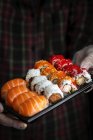 Crop chef irreconhecível em luvas mostrando platter com conjunto de sushi sortido palatável no quarto escuro — Fotografia de Stock