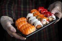 Crop unrecognizable chef in gloves showing platter with set of palatable assorted sushi in dark room — Stock Photo
