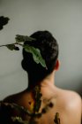 Вид сзади анонимный мужчина без рубашки сидит в темной комнате рядом с нежной тонкой веткой растения с засохшими листьями — стоковое фото