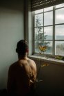 Back view anonymous shirtless male sitting in dark room near tender thin plant twig with withering leaves and looking out window — Stock Photo