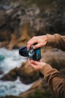Ritaglia anonimo fotografo maschio scattare foto su macchina fotografica professionale di acqua di mare schiumosa lavaggio ruvida scogliere rocciose — Foto stock