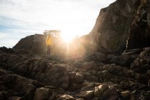 Touriste sans visage de pleine longueur en veste chaude debout sur une pente rocheuse rugueuse pierreuse contre le rayon de soleil et bénéficiant d'une vue sur les hauts plateaux — Photo de stock