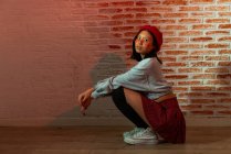 Comprimento total confiante jovem asiático feminino em roupa elegante e chapéu sentado em parquet e olhando para a câmera no quarto escuro contra a parede de tijolo — Fotografia de Stock