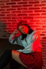 Fiducioso giovane donna asiatica in abito elegante e cappello seduto sul parquet e guardando la fotocamera in camera oscura contro muro di mattoni — Foto stock
