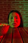 Joven hembra asiática sin emociones con jeroglíficos pintados en una cara bonita apoyada en una mesa de madera con fideos en un tazón y mirando a la cámara - foto de stock