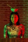 Emotionless young Asian female with painted hieroglyphs on face wearing stylish wear with bowl of noodles on head touching chin gently and looking at camera in dark room — Stock Photo