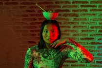 Jeune femme asiatique immobile avec hiéroglyphes peints sur le visage portant une tenue élégante avec bol de nouilles sur la tête touchant le menton doucement et regardant la caméra dans la pièce sombre — Photo de stock